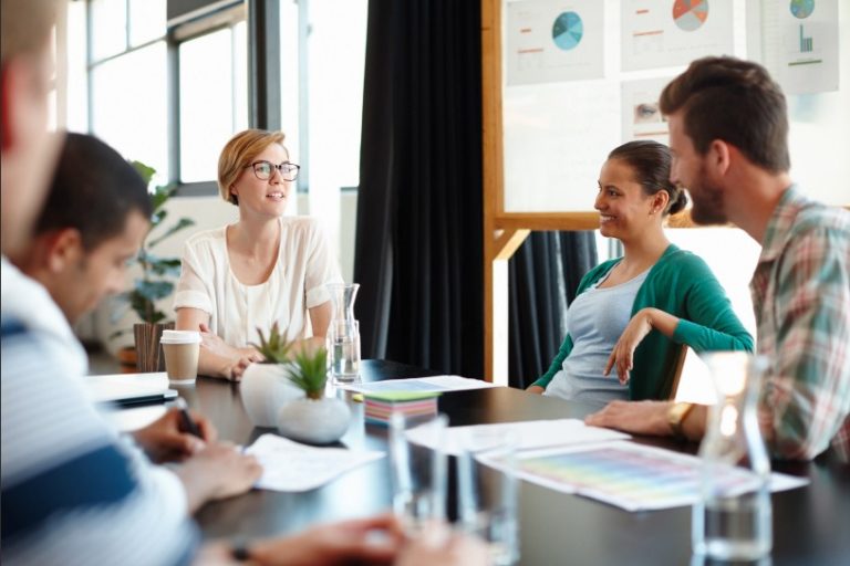 Five young employees in a meeting at a community table. They are smiling and listening to the woman at the head of the table.