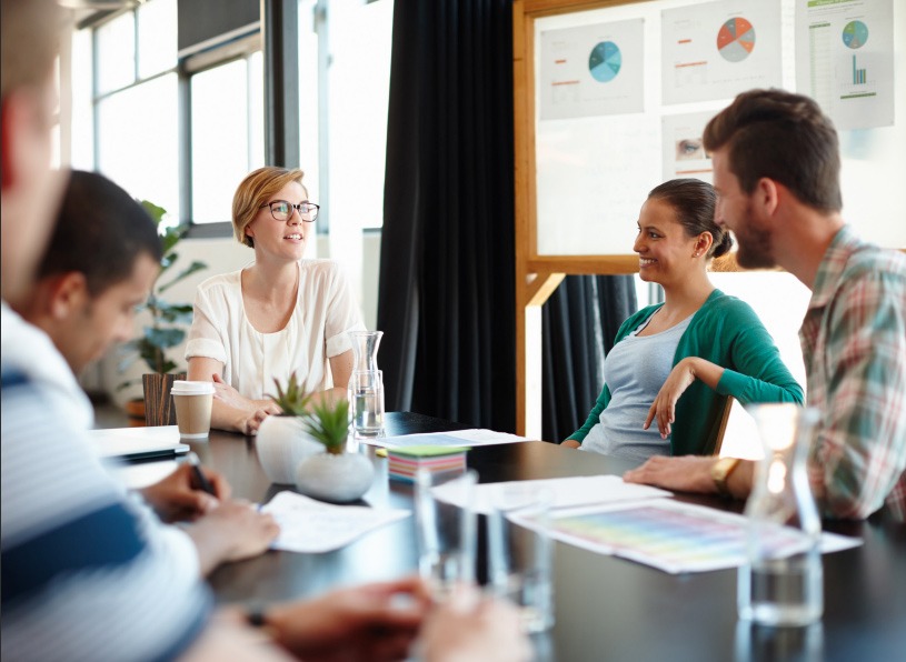 Five young employees in a meeting at a community table. They are smiling and listening to the woman at the head of the table.